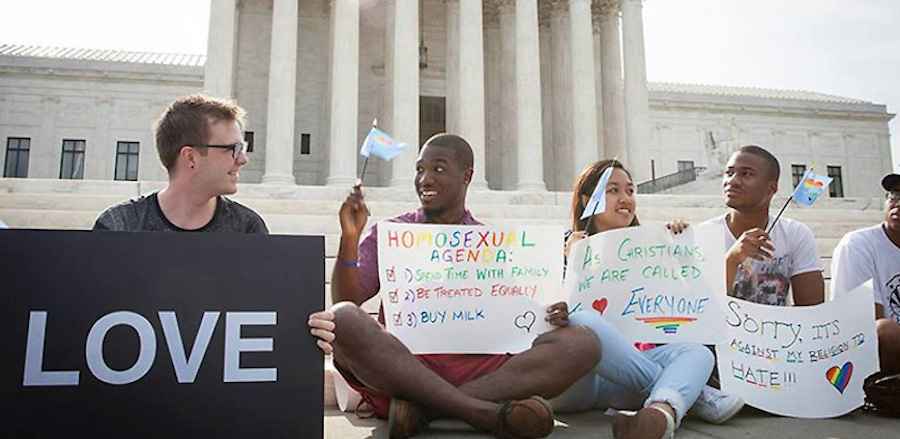 Five people sit on the ground in front of the Capitol holding LGBTQ protest signs and flags 