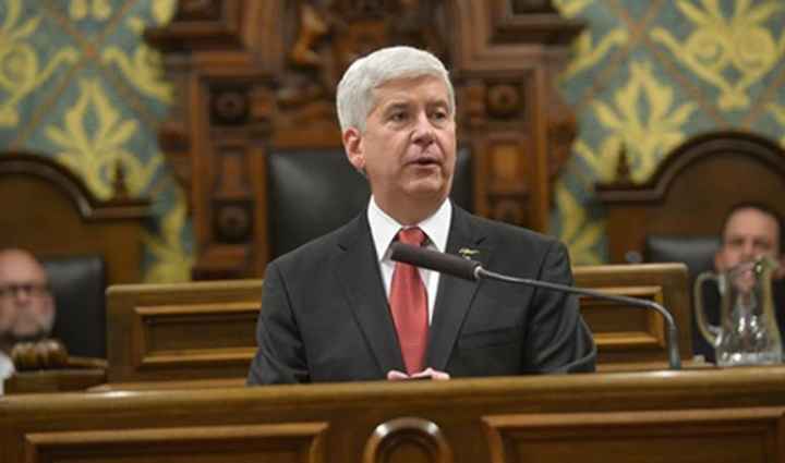 Governor Snyder sits behind a podium speaking into a microphone. He has close cropped grey hair and wears a suit with a red tie. 