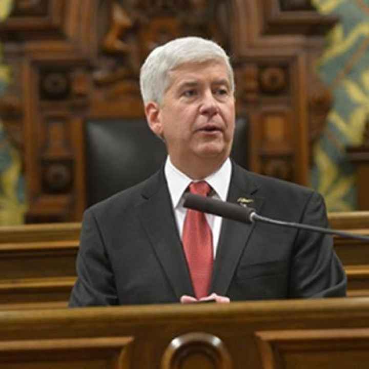 Governor Snyder sits behind a podium speaking into a microphone. He has close cropped grey hair and wears a suit with a red tie. 