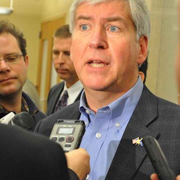 Reporters interview a man who looks stressed. He looks off into the distance as he speaks. He wears a checked suit jacket and blue collar. 