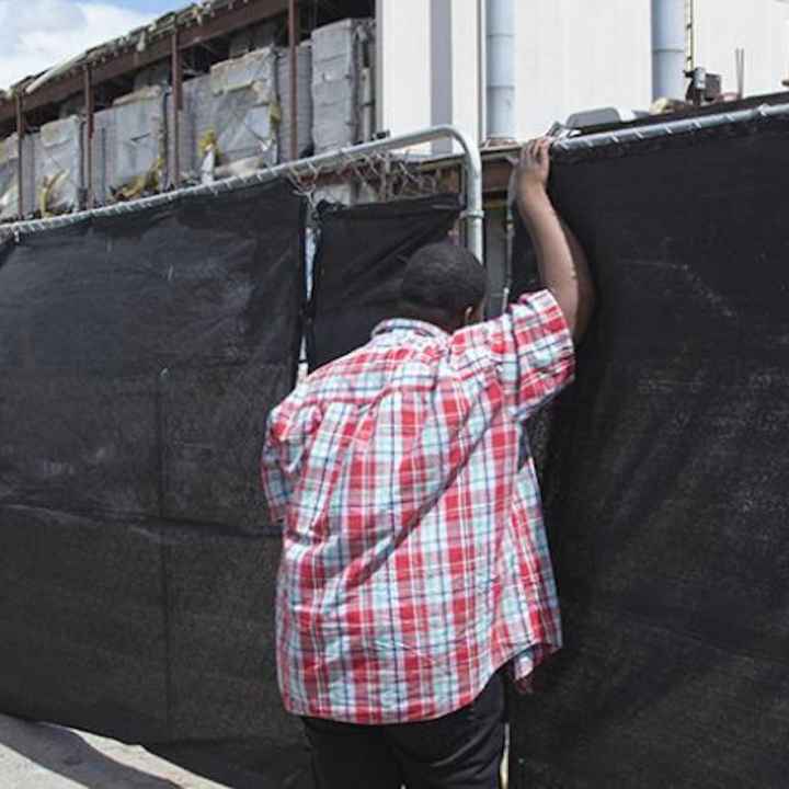 A young boy leans against a fence covered by black cloth. Behind the fence is a construction zone. 