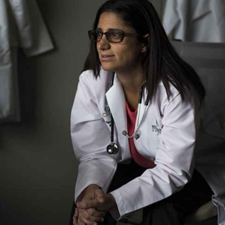 Dr. Mona Hanna-Attisha sits in a chair leaning forward and clasping her hands. She wears black rimmed glasses, a white coat, pink top and dark pants. She looks to the viewer&#039;s left. 