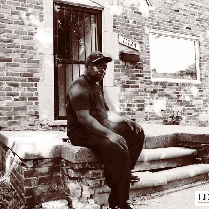 Walter Hicks sits on his home's stopp. He looks at the camera with his hands on his knees, one foot propped on a step.